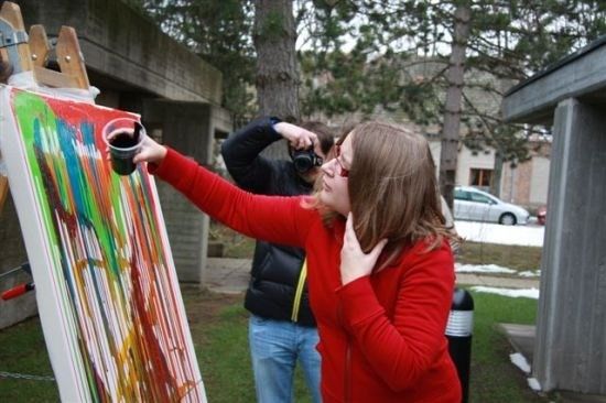 Eine Frau in einem roten Pullover gießt bunte Farbe auf eine Leinwand, während ein Mann hinter ihr ein Foto macht.