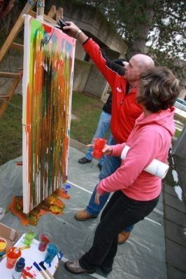 Ein Mann und eine Frau malen ein großes Leinwand im Freien. Der Mann benutzt einen Pinsel, während die Frau einen Becher hält. Verschiedene Farben sind auf dem Boden.