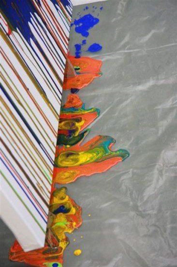 Ein Nahaufnahme einer weißen Oberfläche mit leuchtenden, tropfenden Farben in verschiedenen Tönen wie Blau, Orange und Gelb. Die Farbe läuft auf eine graue, strukturierte Oberfläche.