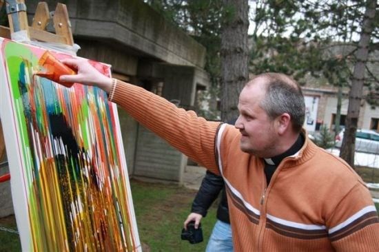 Ein Mann in einem orangefarbenen Pullover malt ein farbenfrohes abstraktes Gemälde auf einer Leinwand im Freien.
