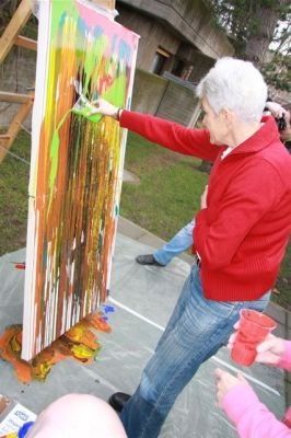 Eine ältere Frau in einem roten Pullover und Jeans malt auf einer großen Leinwand mit leuchtenden Farben. Sie hält einen Pinsel und einen roten Becher. Eine Person in einem rosa Shirt steht in der Nähe.