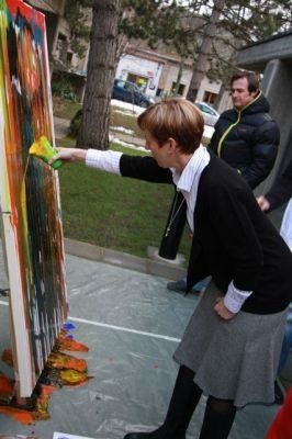 Eine Frau malt ein großes, buntes Bild im Freien, während ein Mann im Hintergrund zusieht.