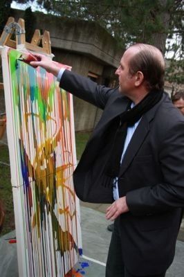 Ein Mann in einem schwarzen Anzug und Schal malt ein farbenfrohes abstraktes Werk auf eine große Leinwand.