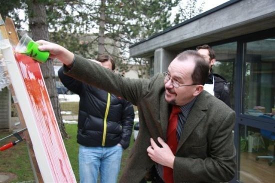 Ein Mann in Anzug und Brille malt ein rotes Bild auf eine Leinwand, während andere zusehen.