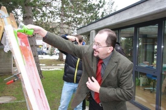 Ein Mann in Anzug und Krawatte malt ein Leinwandbild im Freien. Ein anderer Mann in einer schwarzen Jacke macht ein Foto.