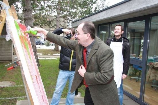 Ein Mann in Anzug und Krawatte sprüht Farbe auf eine große Leinwand, während zwei andere zusehen. Einer hält eine Kamera, und ein anderer trägt eine Schürze.