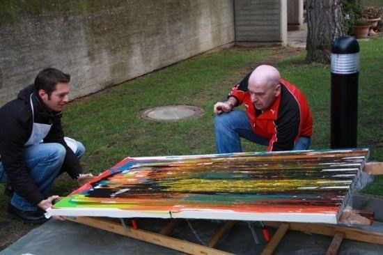 Zwei Männer arbeiten in einem Hinterhof an einem großen, farbenfrohen Gemälde auf einem Holz-Easel.