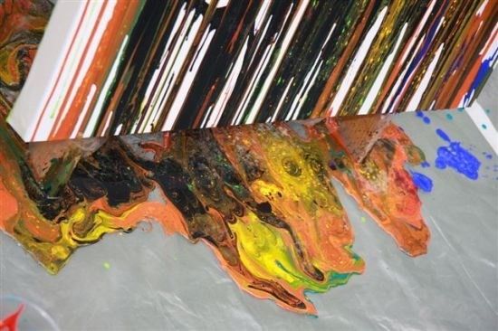 Ein lebendiges abstraktes Gemälde mit orangefarbenen, gelben, grünen und blauen Farben, die auf einem weißen Leinwand auf einer grauen Oberfläche fließen.