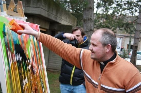 Ein Mann in einem braunen Pullover malt auf einer Leinwand mit einem Pinsel. Ein anderer Mann steht hinter ihm und hält eine Kamera.