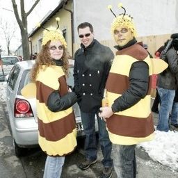 Drei Menschen in Bienenkostümen stehen auf einer verschneiten Straße, mit einem Auto und einem Gebäude im Hintergrund.