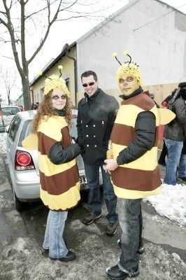 Drei Menschen in Bienenkostümen stehen auf einer verschneiten Straße, mit einem Auto und einem Gebäude im Hintergrund.