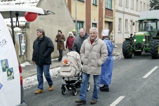 Eine Gruppe von Menschen geht im Winter eine Straße entlang, ein Mann schiebt einen Kinderwagen mit einem Baby, während ein Traktor in der Nähe geparkt ist.