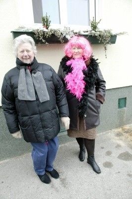 Zwei Frauen, die warm gekleidet sind, eine mit rosa Haaren und einem rosa Boa, stehen vor einem Gebäude.
