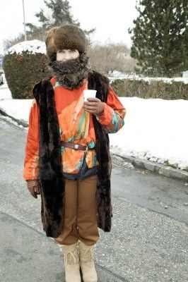 Eine Person in einem Pelzmantel und Bart steht auf einer verschneiten Straße und hält einen Becher.