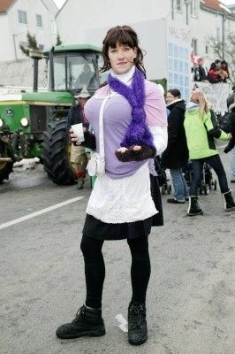 Eine Frau in einem lila und weißen Kostüm mit einem lila Schal steht auf der Straße, einen Becher in der Hand, mit einem Traktor und Menschen im Hintergrund.