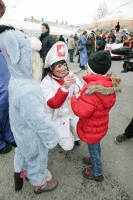 Eine Frau in einer Krankenschwester-Verkleidung interagiert mit einem Kind in einer roten Jacke, während eine weitere Person in einem blauen Tierkostüm in der Nähe steht. Die Szene spielt sich draußen ab, mit mehreren Personen im Hintergrund.