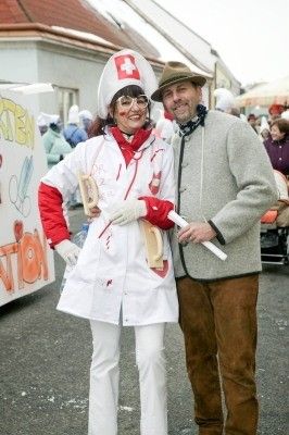 Eine Frau in einer verbluteten Arztuniform und ein Mann in einem grauen Mantel stehen gemeinsam auf einer Straße und lächeln für ein Foto. Hinter ihnen ist ein Banner mit rotem Text zu sehen, und mehrere Personen stehen im Hintergrund.
