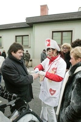 Eine Gruppe von Menschen vor einem Gebäude, eine Frau in einer Krankenschwesteruniform mit einem roten Kreuz auf ihrer Mütze gibt einem Mann ein Glas Bier.