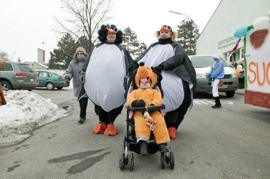 Drei als Pinguine verkleidete Personen, zwei Erwachsene und ein Kind im Kinderwagen, gehen in einem verschneiten Bereich, hinter ihnen sind Autos geparkt.