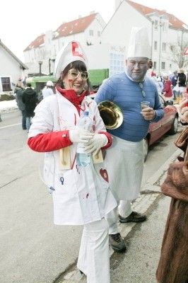 Eine Frau in einer Krankenschwester-Verkleidung und ein Mann in einer Kochmütze stehen am Straßenrand, mit einem roten Auto und Gebäuden im Hintergrund.