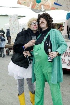 Zwei Personen in Kostümen, eine in einem schwarzen Pelzmantel und die andere in grüner Latzhose, stehen zusammen und posieren für ein Foto. Sie halten Getränke in den Händen und scheinen sich auf einem Outdoor-Event zu befinden.