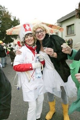Zwei Frauen in weißen Kostümen, eine mit einer Krankenschwestermütze und Brille, die andere mit einem schwarzen Mantel und gelben Stiefeln, posieren für ein Foto auf der Straße.