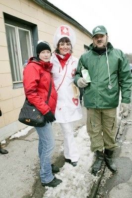Drei Personen in Winterkleidung, eine in einer roten Jacke, eine andere in einer weißen Krankenschwester-Kostüm und ein Mann in einer grünen Jacke, posieren für ein Foto vor einem Gebäude mit Schnee auf dem Boden.