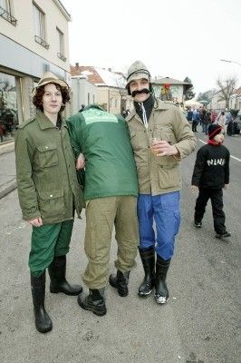 Drei Personen in Militäruniformen und Stiefeln stehen auf einer Straße, eine Person hält ein Glas. Ein Kind läuft hinter ihnen.