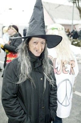 Eine Frau in einer Hexenverkleidung mit schwarzem Hut und langen grauen Haaren steht im Freien, lächelt und posiert für ein Foto. Hinter ihr ist eine weitere Person mit einem Friedenszeichen-Shirt zu sehen.