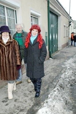 Drei Personen gehen auf einer verschneiten Straße vor einem Gebäude mit einer grünen Tür. Die Frau links trägt einen Pelzmantel und Stiefel, der Mann in der Mitte einen grünen Schal und die Frau rechts hat rote Haare und einen schwarzen Mantel.
