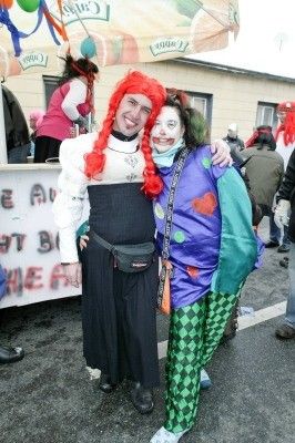 Zwei Personen in farbenfrohen Kostümen, eine mit roten Haaren und die andere in einem mehrfarbigen Outfit, stehen zusammen im Freien. Hinter ihnen ist ein Imbissstand mit Luftballons und einem Gebäude zu sehen.