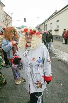 Eine Frau mit blonden Locken trägt eine Blumengirlande und einen bunten Mantel mit Aufnähern auf der Straße während eines Umzugs.