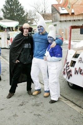 Drei Menschen in Kostümen, einer mit blauer Gesichtsbemalung, stehen zusammen auf einer Straße mit einem Auto und Gebäude im Hintergrund.