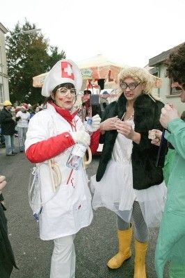 Zwei Frauen in Kostümen, eine als Krankenschwester mit rotem Kreuz, die andere in einem weißen Kleid und gelben Stiefeln, stehen in einem Außenbereich mit anderen Personen in Kostümen.