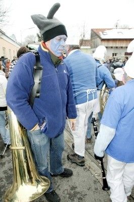 Ein Mann mit blauer Gesichtsbemalung und Kostüm hält eine Tuba und steht auf einer Straße mit anderen Menschen in ähnlichen Kostümen.