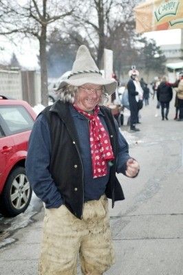 Ein Mann in traditioneller Kleidung mit Hut und Schal steht auf einem Bürgersteig, lächelt und schaut in die Kamera. Hinter ihm steht ein rotes Auto und Menschen gehen auf dem Bürgersteig.