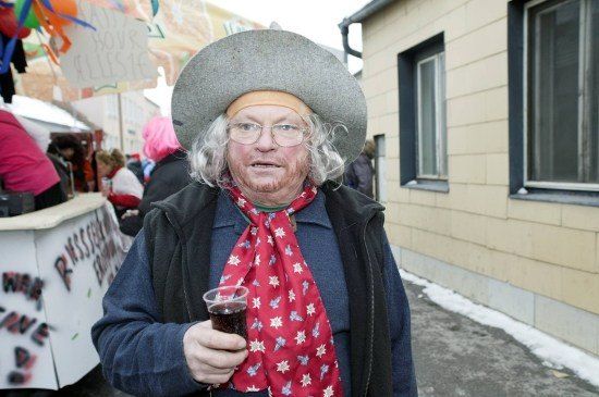 Ein älterer Mann in einem Cowboyhut und rotem Schal hält ein Getränk vor einem Gebäude mit verschneitem Bürgersteig.
