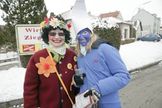 Zwei Frauen in farbenfrohen Kostümen, eine mit blauem Gesicht und die andere mit einer Blumenkrone, stehen auf einer verschneiten Straße mit einem Schild, das 'Wirt Zieg' im Hintergrund steht.