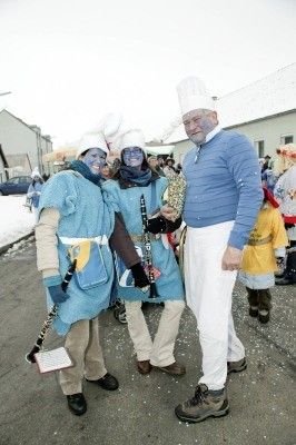 Drei Personen in blauen Kostümen mit Kochmützen und Musikinstrumenten posieren für ein Foto auf einer verschneiten Straße.