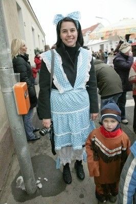 Eine Frau in einem gepunkteten Kleid und ein Kind in einer Mütze stehen auf einer Straße, mit einer Menge Leute im Hintergrund.