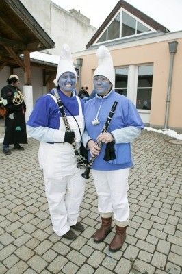 Zwei Personen in blauen und weißen Kostümen mit blauer Gesichtsbemalung, die Klarinetten halten, stehen auf einem Ziegelweg vor einem Gebäude.