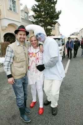 Drei Menschen in Kostümen stehen auf einer verschneiten Straße, einer mit blauer Gesichtsbemalung und weißer Kleidung, ein weiterer mit Hut und Stiefeln und ein dritter in einem karierten Hemd.