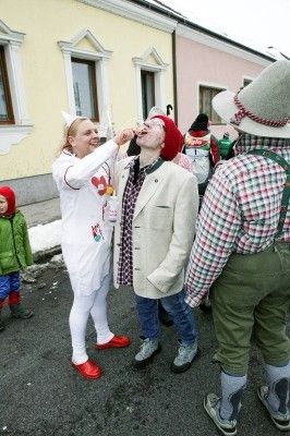 Eine Frau in einem weißen Kostüm mit roten Schuhen füttert einen Mann in einem Anzug und rotem Hut, während ein anderer Mann in traditioneller Kleidung in der Nähe steht, alles in einer festlichen Außenumgebung.