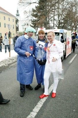 Drei Menschen, die als medizinisches Personal gekleidet sind, stehen lächelnd und posieren für ein Foto auf der Straße. Sie stehen vor einem weißen Van mit einem roten Kreuz. Der Hintergrund zeigt eine verschneite Straße mit Fußgängern und einem Gebäude.