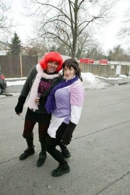 Zwei Frauen in farbenfrohen Kostümen, eine mit roten Haaren, stehen in einem verschneiten Parkplatz mit einem Holzzaun und Bäumen im Hintergrund.