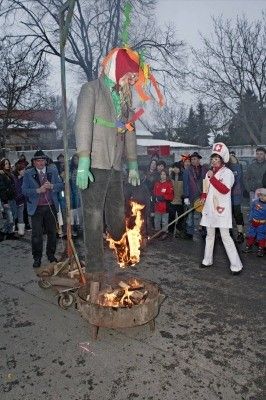 Eine große Puppe wird auf einem öffentlichen Platz verbrannt, während eine Menschenmenge zuschaut. Die Puppe ist in ein farbenfrohes Kostüm gekleidet und hat ein Feuer an ihren Füßen. Eine Person in einem weißen Mantel steht in der Nähe und hält einen Stock.