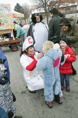 Eine Frau in Arztkluft untersucht ein Kind in einem blauen Elefantenkostüm, während ein weiteres Kind in einer roten Jacke zuschaut, mit einem Pinguin-Maskottchen in der Nähe und einem Imbisswagen im Hintergrund.