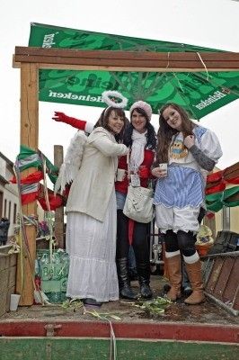Drei Frauen in festlichen Kostümen, eine mit Engelsflügeln, posieren für ein Foto auf einer dekorierten Bühne mit einem grünen Baldachin.