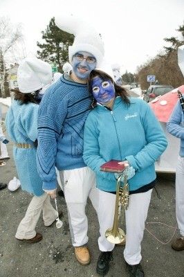 Ein Paar in blauen Kostümen und Gesichtsbemalung, das nebeneinander steht und eine Trompete hält, mit anderen Personen in ähnlichen Kostümen dahinter.