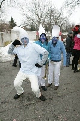 Drei Personen in weißen Kochkostümen mit blauer Gesichtsbemalung, eine hält eine Trompete, stehen auf einer Straße mit Schnee am Boden und einer Wand im Hintergrund.